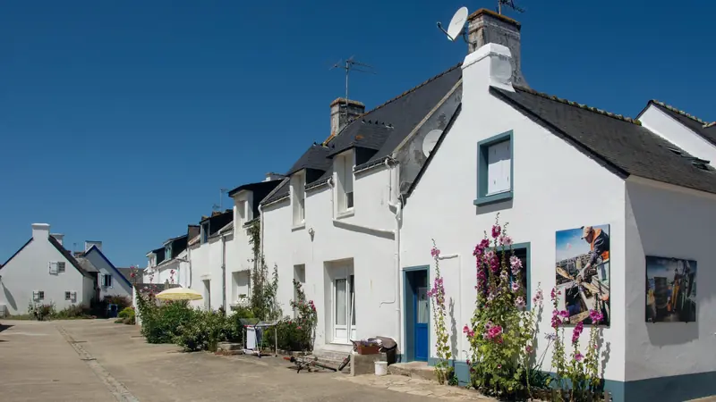 Rue de village breton bordée de maisons blanches aux toits d'ardoise, aux volets bleus et aux roses trémières en fleurs