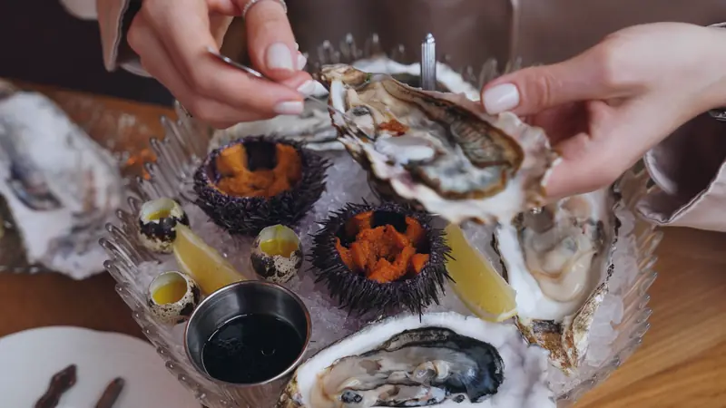 Mains tenant une huître ouverte au-dessus d'un plateau de fruits de mer sur glace, avec oursins et quartiers de citron