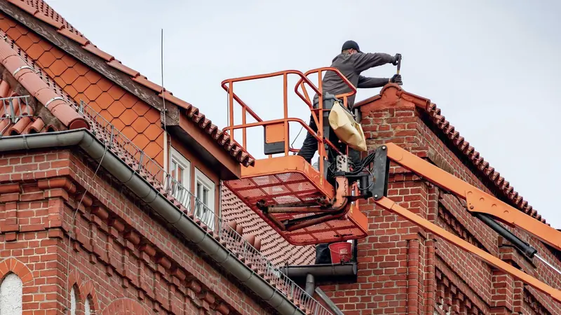 Artisan sur une nacelle réparant le haut d'un pignon en brique sous un toit de tuiles