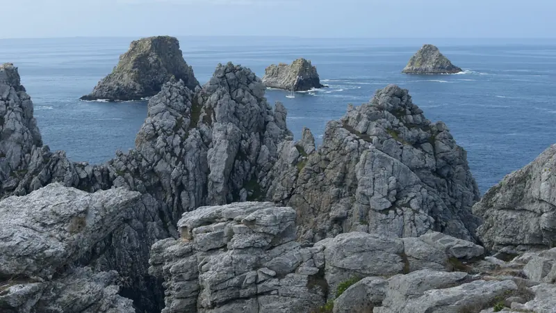 Aiguilles de roche grise et îlots au large d'une pointe bretonne, mer calme et voilier au loin