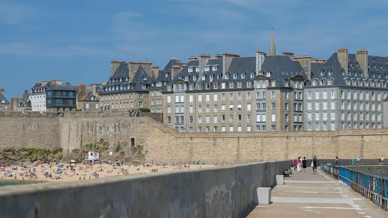 Remparts et immeubles de granit de l'intra-muros de Saint-Malo au-dessus d'une plage fréquentée, vus depuis la jetée