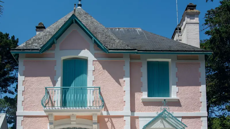 Façade rose d'une villa balnéaire ancienne aux volets turquoise, avec balcon en fer forgé et toit d'ardoise