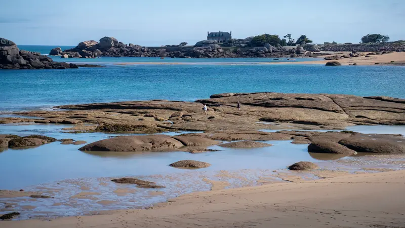Côte de granit rose à marée basse, avec des dalles rocheuses découvertes, des flaques et du sable au premier plan
