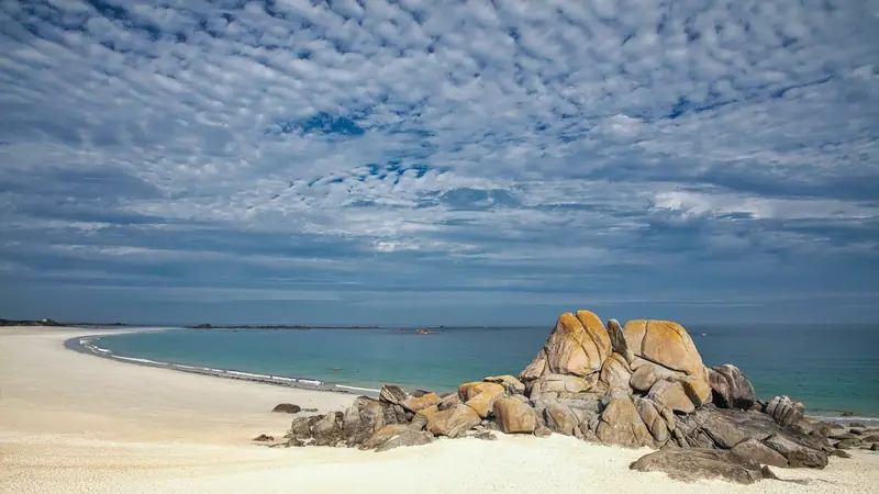 Grande plage de sable blanc en arc de cercle, eau turquoise et amas de rochers de granit sous un ciel moutonné, en Bretagne