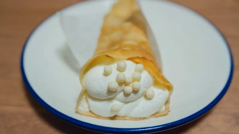 Crêpe fine roulée en cornet, garnie de crème fouettée et de perles de chocolat blanc, sur une assiette à liseré bleu