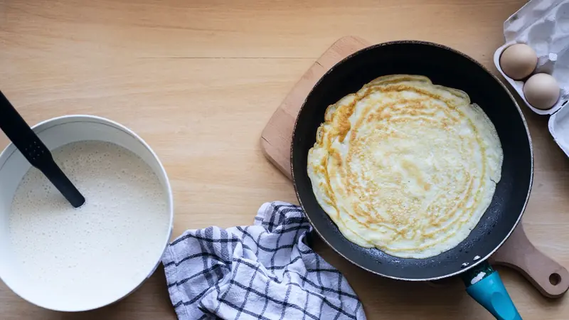 Saladier de pâte à crêpes et crêpe dorée en cuisson dans une poêle, œufs dans leur boîte à côté