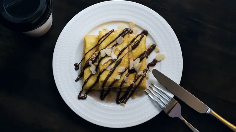 Crêpes pliées nappées de filets de chocolat et d'amandes effilées sur une assiette blanche, gobelet de café à côté