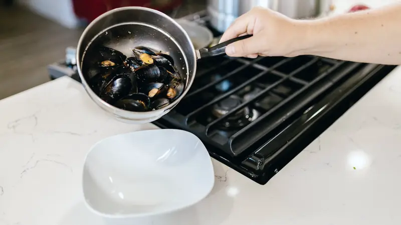 Main versant des moules cuites d'une casserole vers une assiette creuse, près d'une plaque de cuisson