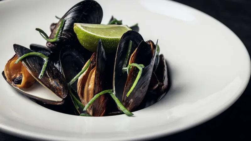 Moules cuites ouvertes dans une assiette creuse blanche, avec un quartier de citron vert et des lanières de poireau