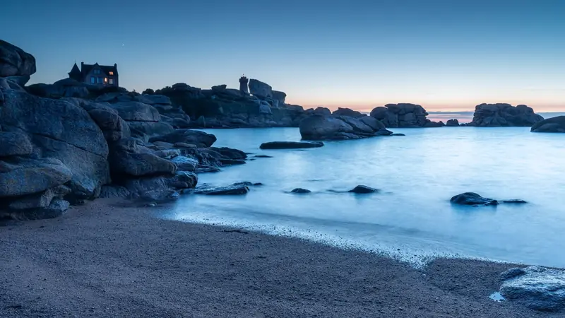 Maison éclairée au-dessus d'une crique de granit rose à la tombée de la nuit, avec un phare au loin